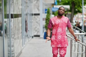 A handsome Ghanaian decent African man posing for the camera and wearing a bright coloured pink shirt and short made with Kente cloth. 